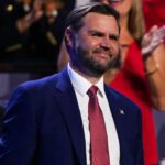 A man in a blue suit and red tie smiles while standing at an event. People are visible in the blurred background, including someone in a white USA hat.