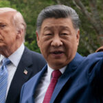 Donald Trump and Xi Jinping in suits stand outdoors; Xi gestures with his hand while the Trump looks on with a serious expression. Trees and greenery are visible in the blurred background.