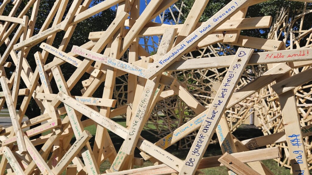 A large, complex sculpture made of interlocking light-colored wooden planks covered in handwritten messages and signatures.