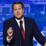 A man in a suit and red tie speaks into a microphone at a podium against a blue background.