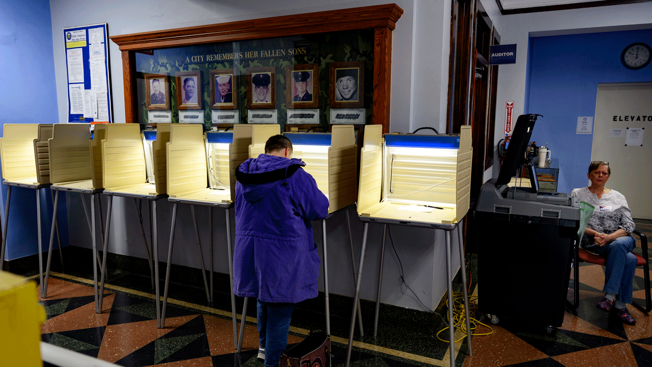 A person in a purple coat fills out a ballot at a voting booth in a room decorated with a memorial wall, while another person sits in a chair nearby.