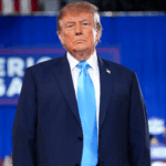 Donald Trump wearing a dark suit and bright blue tie, standing in front of a blue Make America Great Again sign.