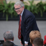 Mike DeWine in a suit and red tie smiles while standing before an audience.