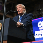 Donald Trump speaking at a podium with a Presidential Seal, in front of a blue screen that reads Ohio Welcomes President Donald J. Trump.