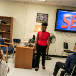 A woman in a red top stands in a classroom in front of a large screen displaying the word SEX in large blue letters as she speaks to a group of seated students.