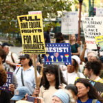 A diverse group of protesters holds signs opposing Immigration and Customs Enforcement, advocating for illegal aliens. Signs include messages like Full and equal rights for all immigrants and Resist! Trees and buildings are visible in the background.