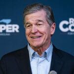 North Carolina Governor Roy Cooper smiling in front of a blue campaign banner amid his Senate race.