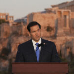 Marco Rubio speaks at a podium with the Parthenon and the Acropolis in the background.