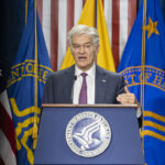 Mehmet Oz in a suit speaking at a podium with the Department of Health and Human Services seal.