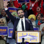 DSA-aligned Zohran Mamdani waves to a cheering crowd while standing at a podium with a sign that reads New York is not for sale.