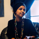 Ilhan Omar wearing a black headscarf and a colorful beaded necklace, sitting in a leather chair.