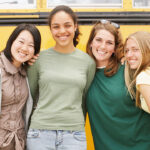 Illinois parents are considering a lawsuit after a school district stood by its policy of placing boys with girls on overnight trips. Four smiling young women standing in front of a yellow school bus.