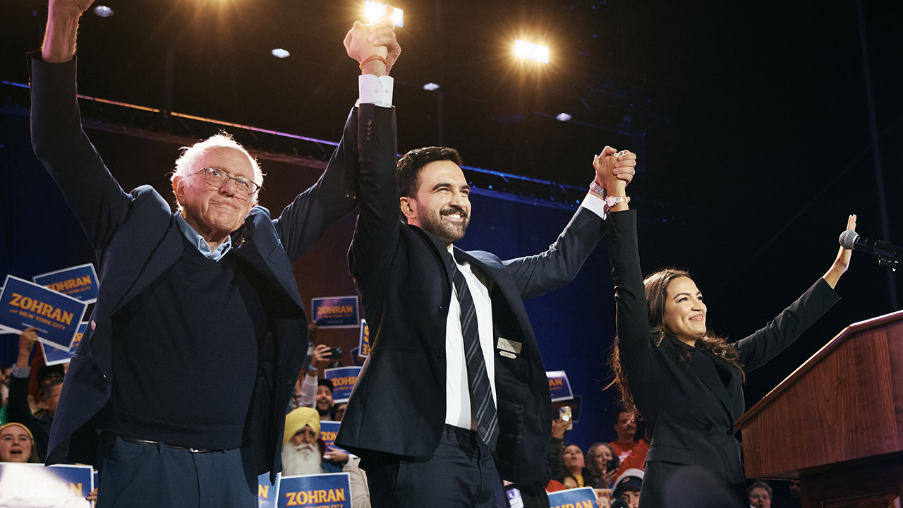 Three major leaders who capture the essence of socialism: Bernie Sanders, Zohran Mamdani, and Alexandria Ocasio-Cortez hold their arms up in celebration behind a podium at a political rally.