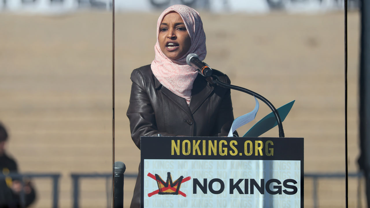 Home 1 Ilhan Omar in a pink headscarf speaks at a NOKINGS.ORG podium, highlighting immigrants’ voices, with NO KINGS and a crossed-out crown graphic. Steps are visible in the background.