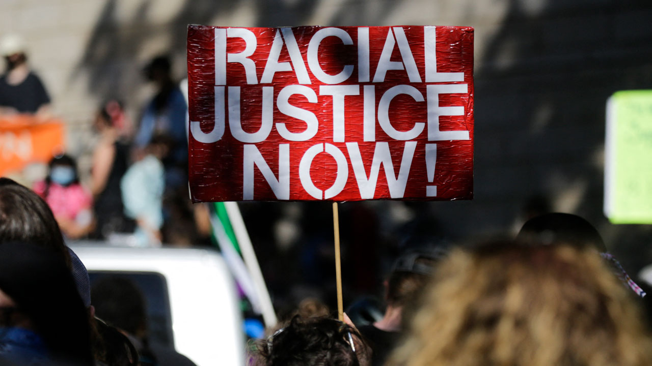 A protestor holds a red and white sign reading RACIAL JUSTICE NOW! in a crowd of people gathered outdoors.