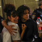 A woman with an emotional expression is comforted by another woman with her arm around her shoulders as a police officer stands nearby.