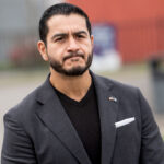 Abdul El-Sayed with a beard, often a target of Islamophobia, wears a dark blazer over a v-neck shirt, with a small flag pin on his lapel.