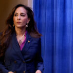 A woman with long dark hair, wearing a navy blue blazer over a purple top and a pearl necklace, stands in front of a blue curtain and a portion of an American flag.