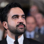 Mamdani with a beard stands before a microphone, speaking to an audience.