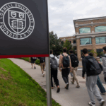Students with backpacks walk along a sidewalk on the Cornell University campus, passing a sign bearing the university seal.
