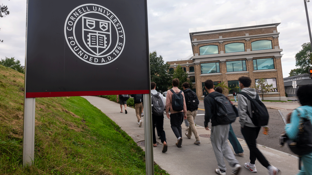 Home 1 Students with backpacks walk along a sidewalk on the Cornell University campus, passing a sign bearing the university seal.