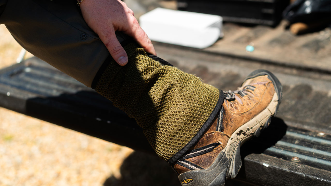 A persons hand adjusts an olive green mesh gaiter worn over a muddy brown hiking boot.