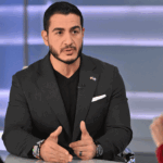 El-Sayed sits at a desk in a black blazer and v-neck shirt during a televised interview, gesturing while speaking to a person in red who's mostly off-camera.