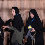 Four women in Iran standing in a line, each holding an assault rifle against a textured, light brown backdrop.