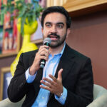 Mamdani, a man with a beard wearing a black blazer and blue dress shirt, speaks into a microphone while gesturing during an event.