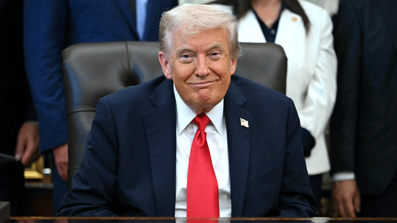 Home 1 Donald Trump in a dark suit and red tie sits at a desk with people standing behind him.
