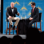 Jeb Bush and Daniel Garza sit on stools on a stage in front of blue The Libre Initiative banners, appearing to participate in a panel discussion.