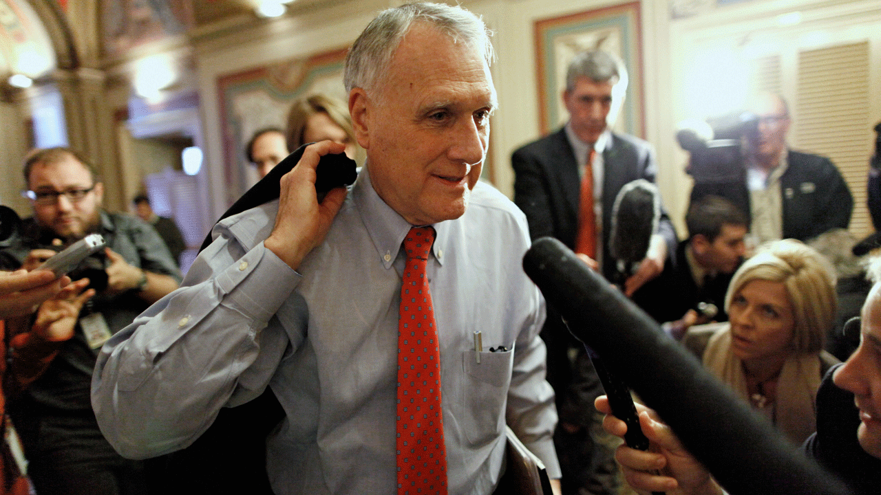 Sen. Jon Kyl walks through a crowd surrounded by reporters holding microphones.
