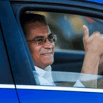 Xavier Becerra, wearing glasses, sits in the driver’s seat of a blue car and gives a thumbs up.