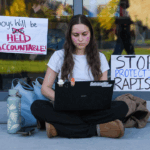 A student sits cross-legged on the ground using a laptop during Cornell 7, flanked by signs reading Boys will be held accountable and Stop protecting rapists.