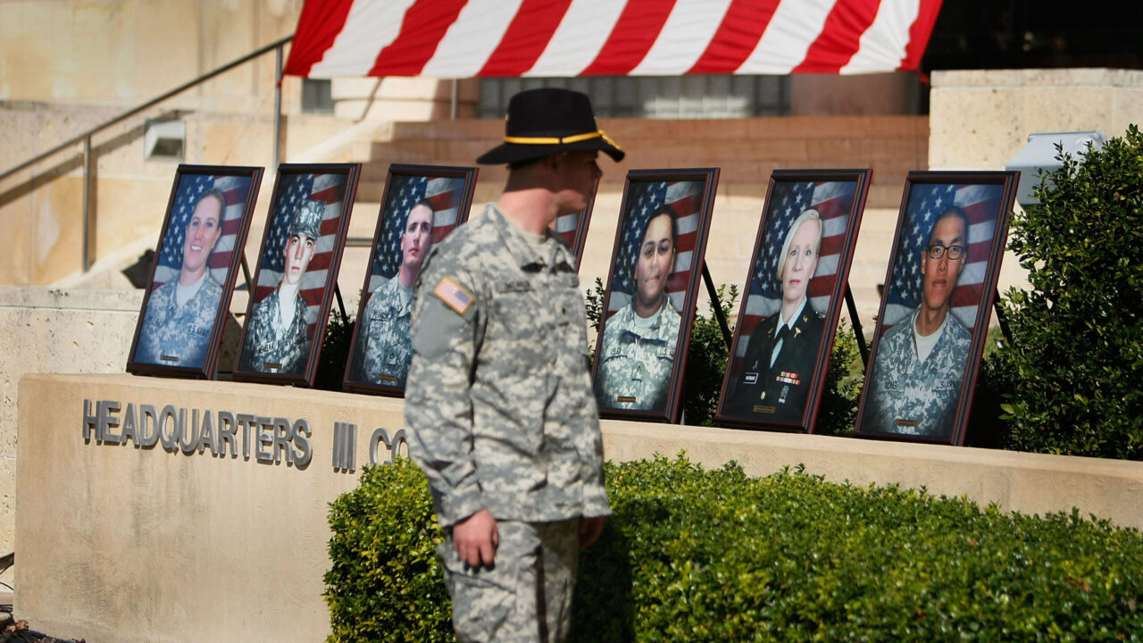 A soldier in uniform stands at Fort Hood in front of a row of framed portraits of service members displayed on a stone wall in front of an American flag.