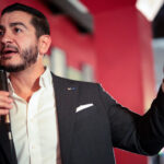 Abdul El-Sayed in a suit jacket and white shirt speaks into a microphone while gesturing with his left hand.