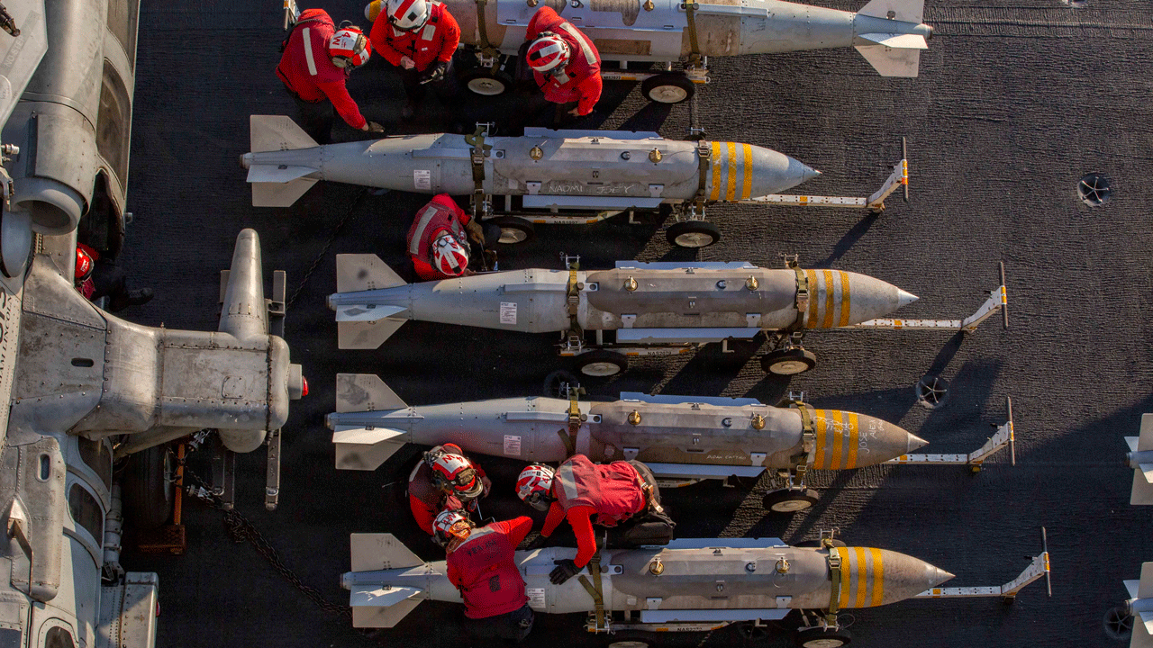 Home 1 Military flight deck crew members in red jerseys prepare and maneuver rows of aerial bombs on the deck of an aircraft carrier.