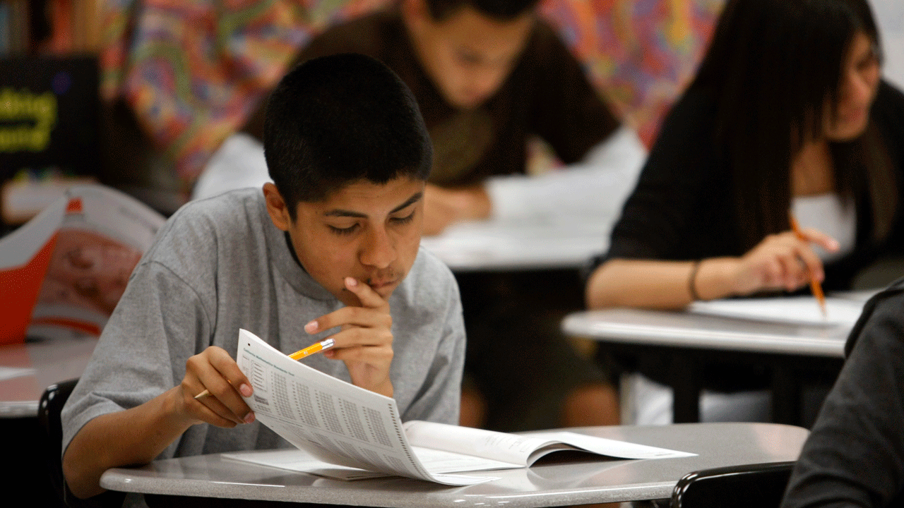 Home 1 A student sits at a desk in a classroom looking down at a test textbook, with other students visible in the background.