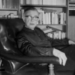 Jean-Paul Sartre, a prominent figure in Europe, wearing glasses and sitting in an armchair in front of a bookshelf.