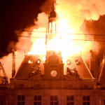 A large building with a clock tower in France engulfed in flames at night.