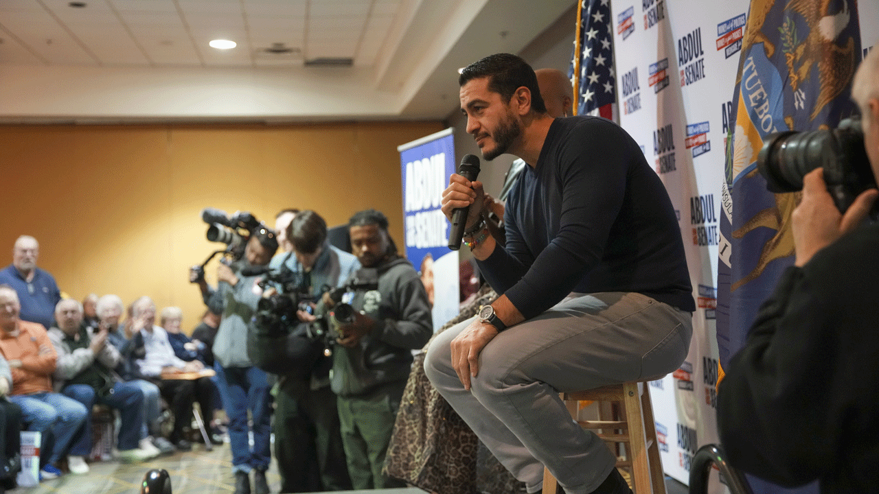 Abdul El-Sayed speaks into a microphone while sitting on a stool at a midterm press conference, surrounded by photographers.