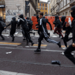 A group of people wearing dark clothing and face coverings, run across a city street with tram tracks.