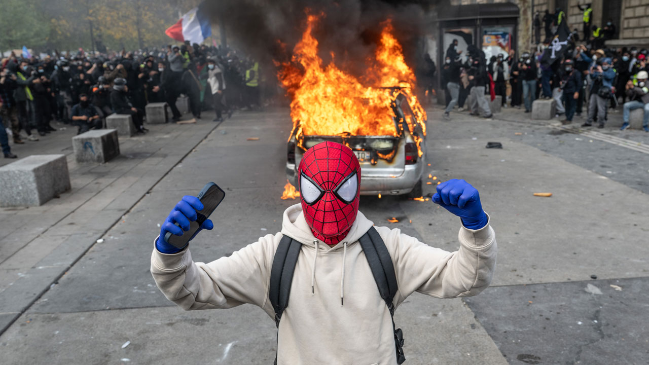 Home 1 A person wearing a Spider-Man mask and blue gloves flexes their arms in front of a car engulfed in flames during a street protest.