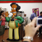 A woman in a pink top waves next to a life-size cutout of Apu from The Simpsons in a convenience store, while another person photographs them.