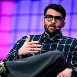 A man with a beard and glasses wearing a dark plaid shirt gestures while sitting on a stage.