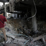 A person stands amidst the ruins of a destroyed building, looking into a large crater in the ground.