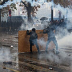Two protesters hold a plywood shield against clouds of smoke in a street lined with police vans.