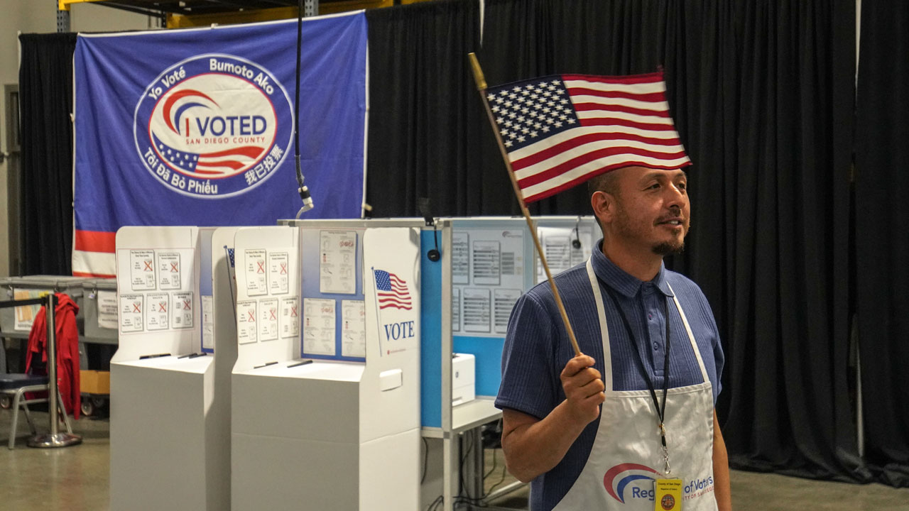 A poll worker wearing an apron and a US flag headband stands in front of voting booths and an I Voted banner during the midterms.