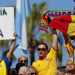 Fans in Romania soccer jerseys hold up a sign reading ROMANIA with a heart and USA, alongside a Romanian flag, in a Lazurcă display of support.