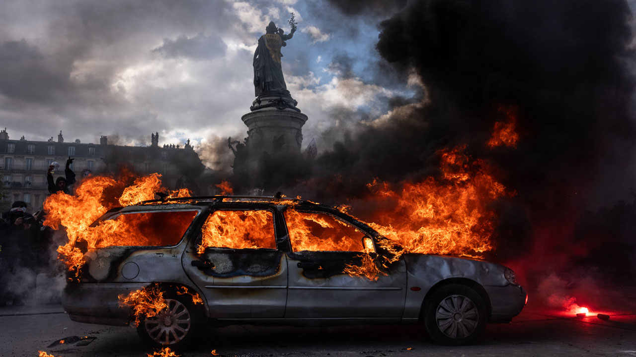 Home 2 A station wagon engulfed in flames in a French city square, with a statue and dark smoke in the background.